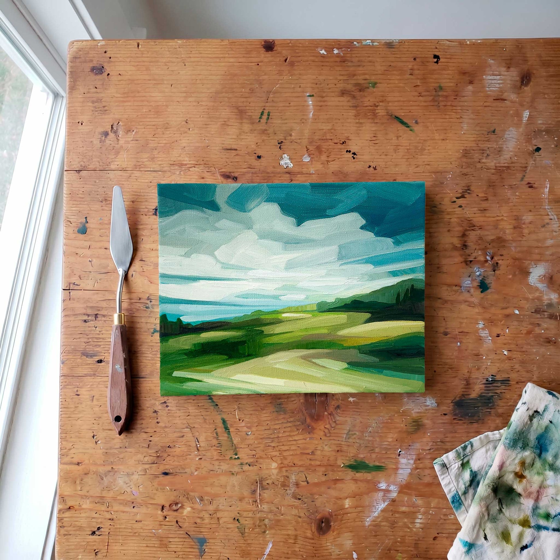 overhead view of an original oil countryside painting with green rolling hils and a blue sky with sweeping clouds