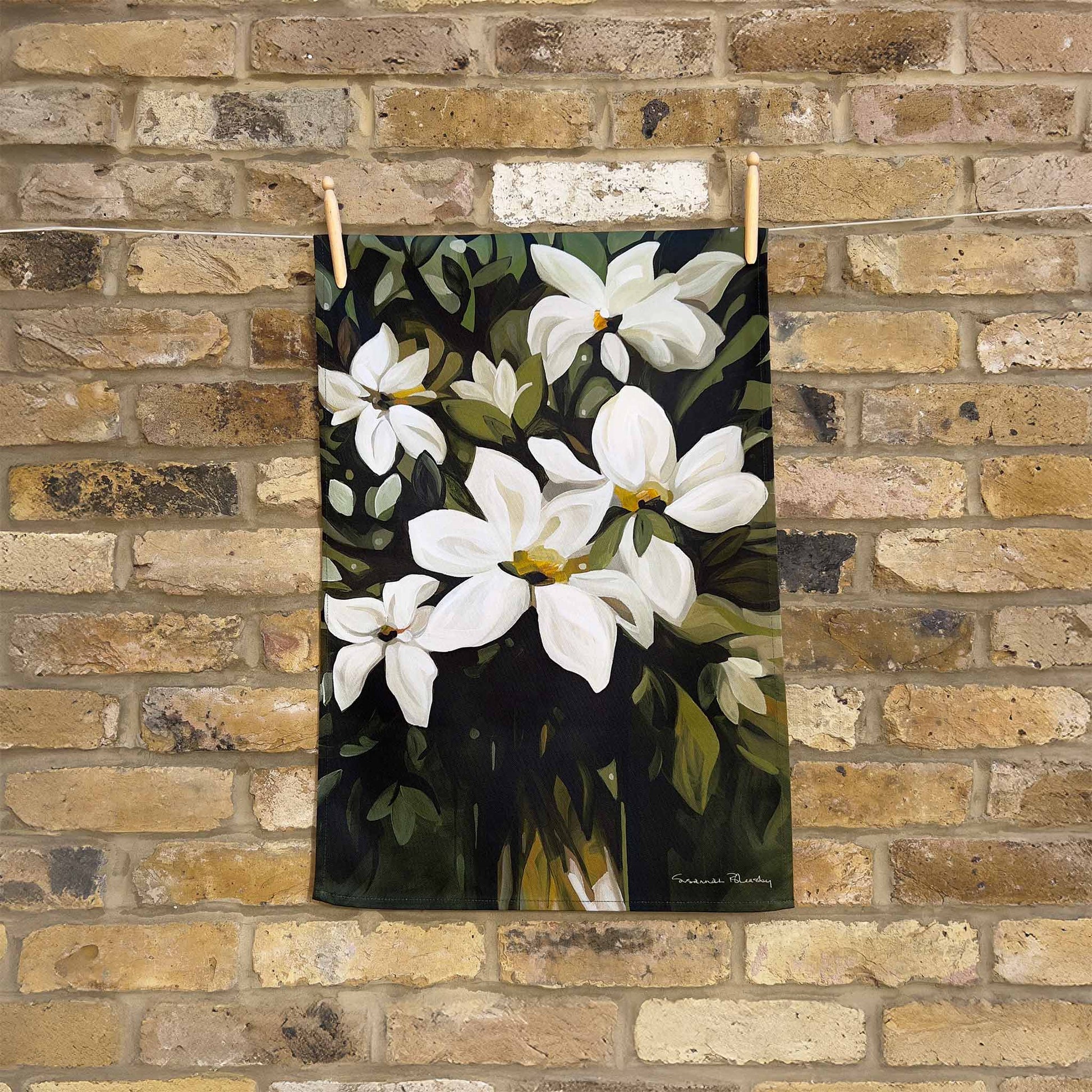 floral cotton tea towel with white flowers on a dark green background hanging against a brick wall
