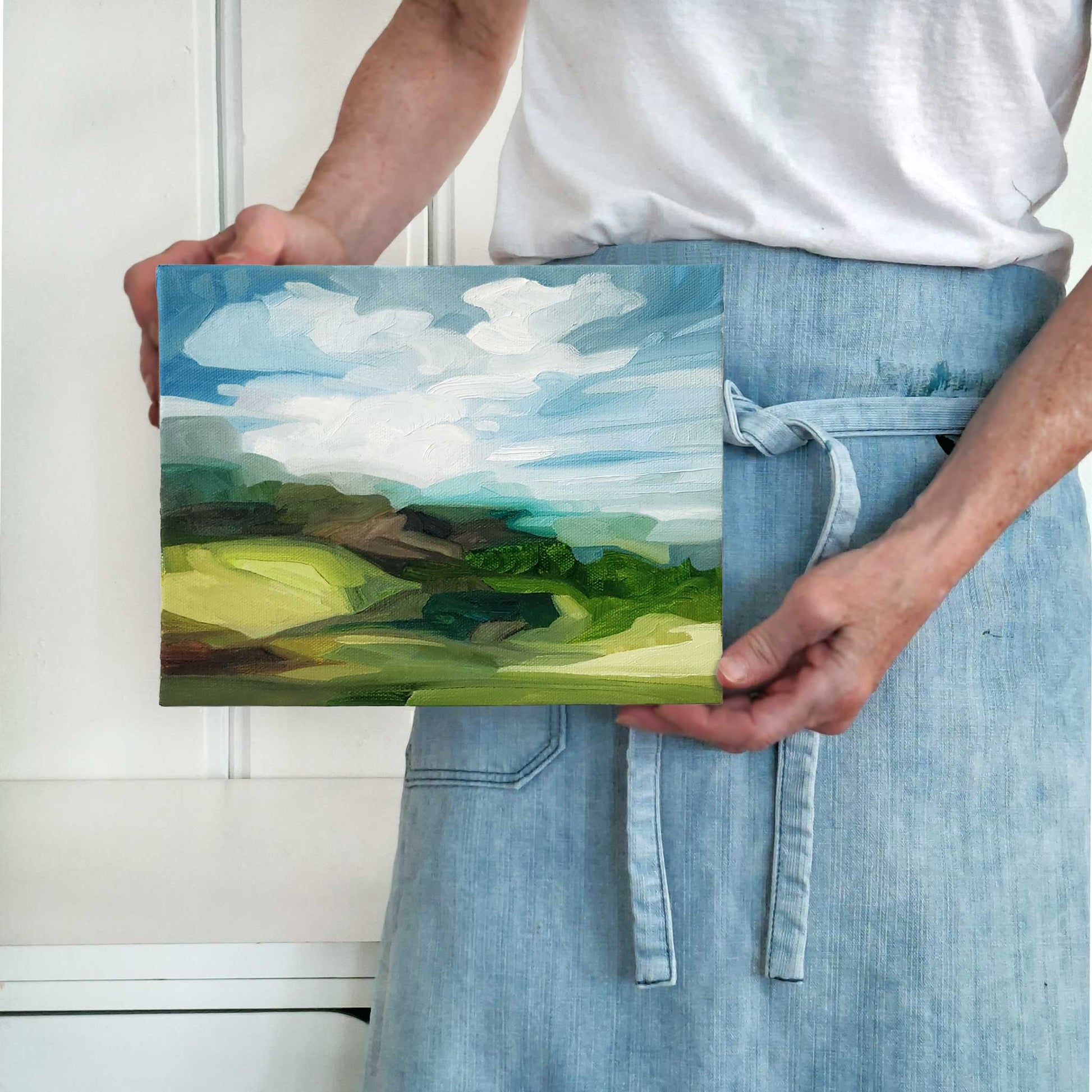 artist holding an original landscape oil painting with blue sky and roling green hills