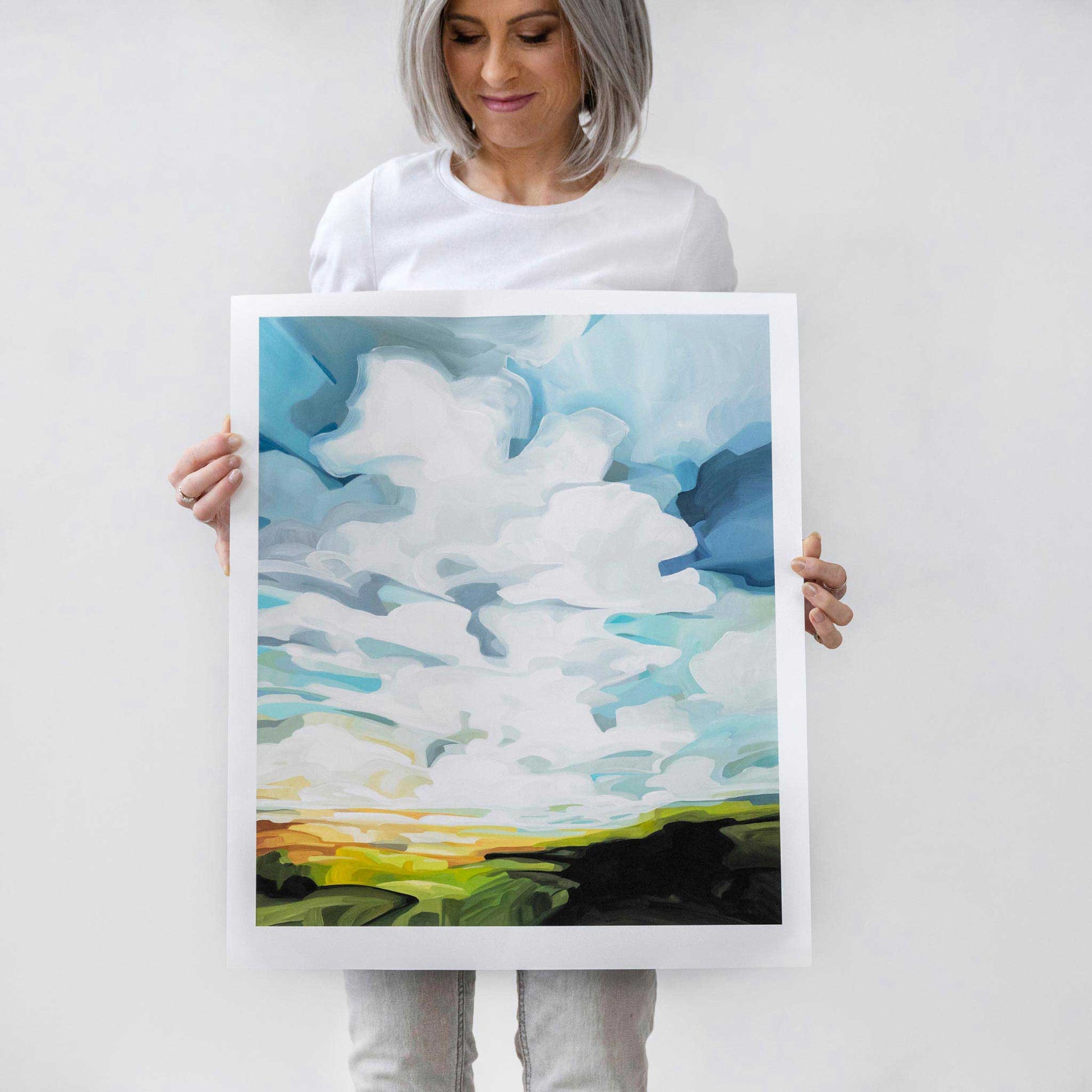 artist holding a limited edition fine art print of an acrylic landscape painting of green hills warm sun and bright blue sky